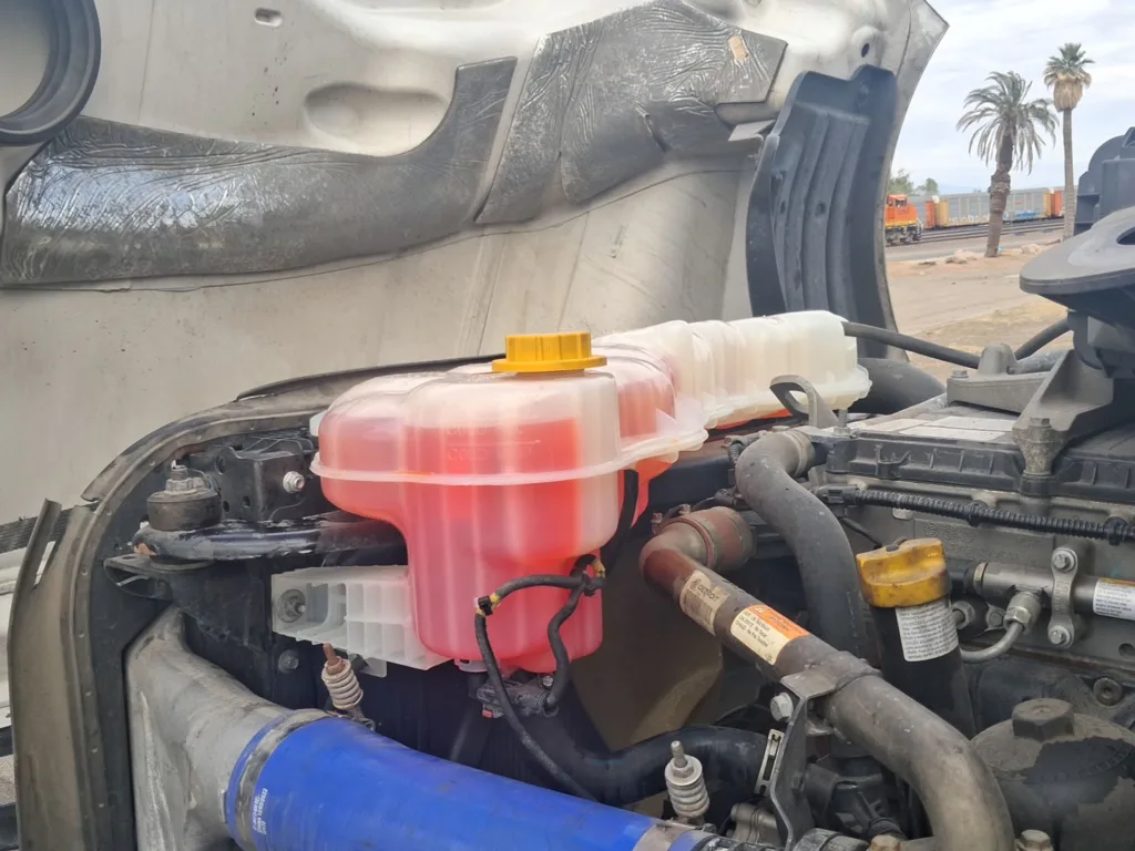 A close-up of a semi-truck engine coolant reservoir with bright red fluid and a yellow cap.