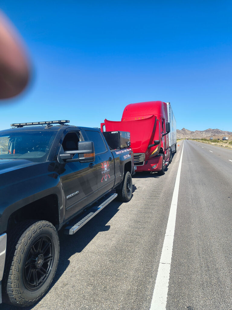Black TRT Roadside Service pickup truck assisting a red semi-truck with its hood open on a highway shoulder.