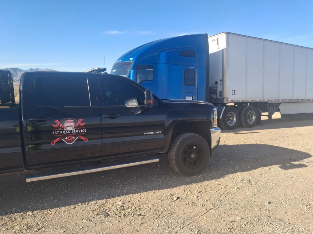 TRT Road Service pickup truck providing 24/7 roadside assistance to a blue semi-truck on a dirt lot.