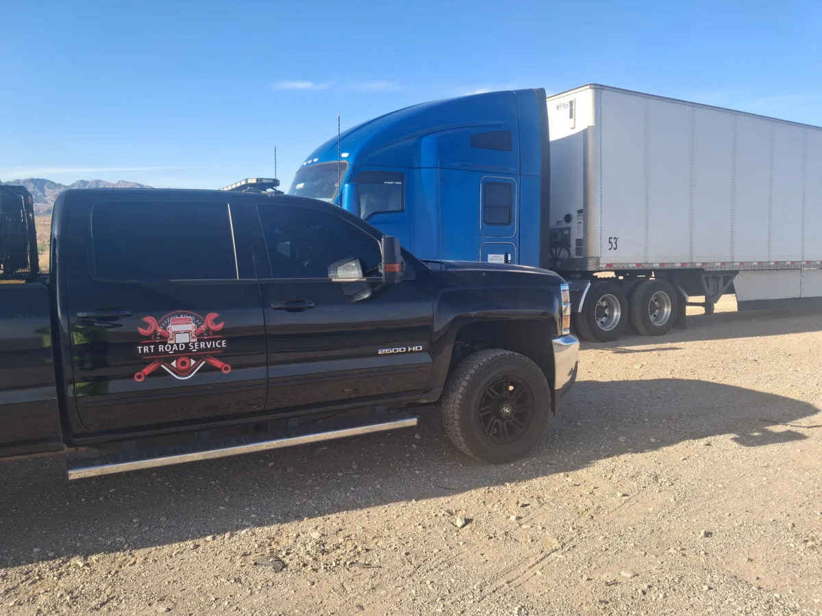 TRT Road Service pickup truck providing 24/7 roadside assistance to a blue semi-truck on a dirt lot.